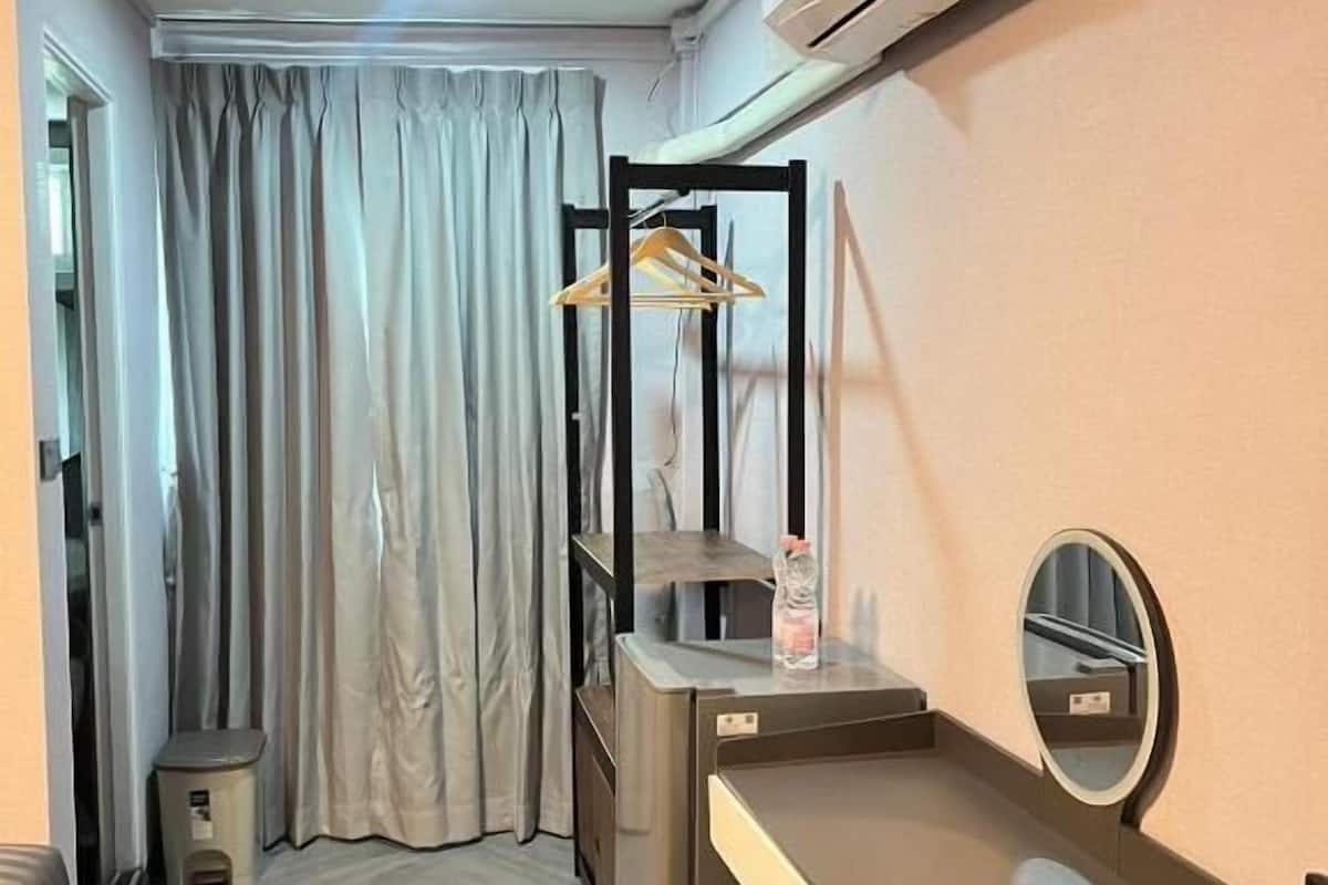 Quarto compacto com cama, escrivaninha com espelho, ar-condicionado, cabideiro e um pequeno banquinho.