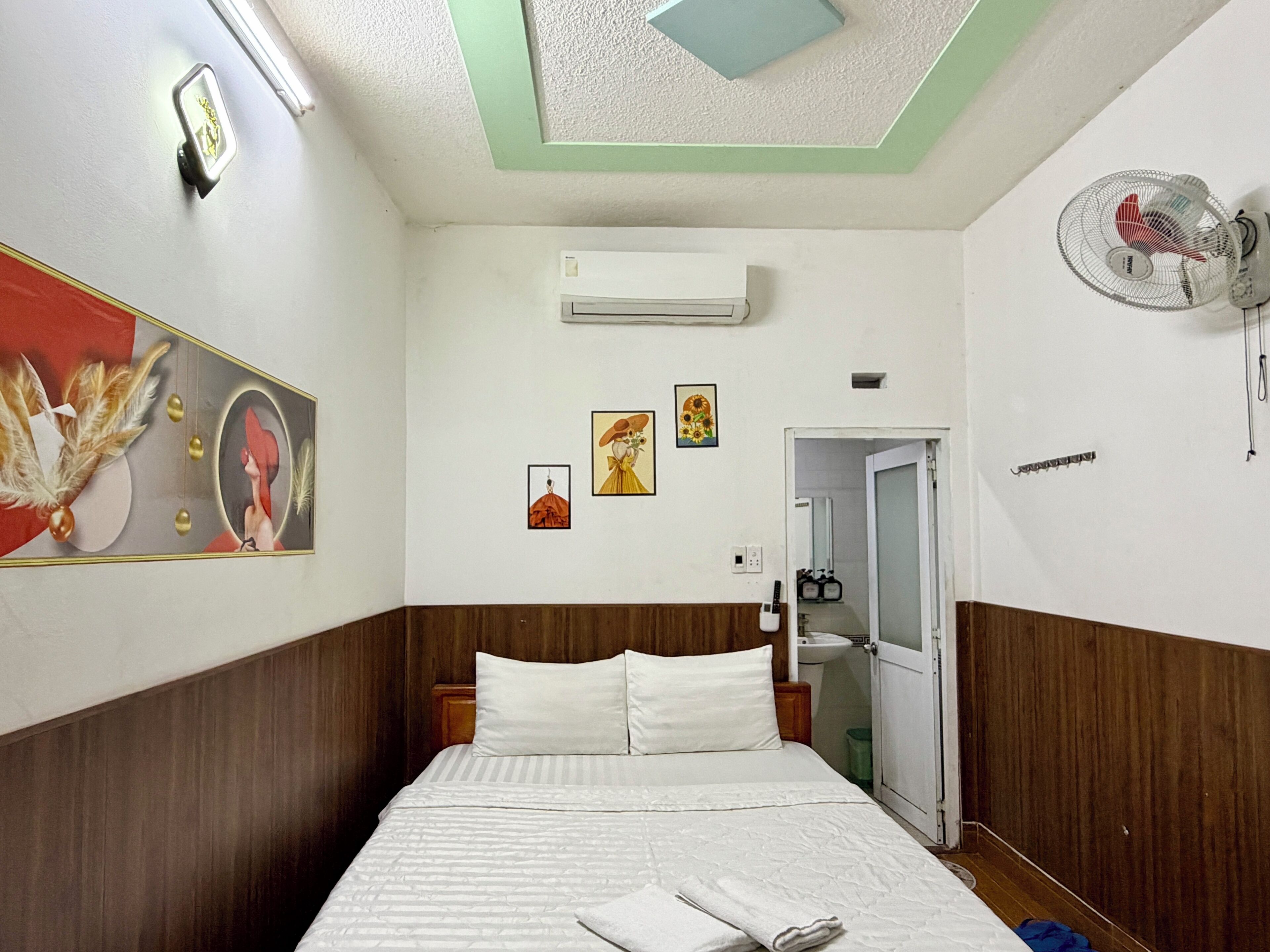 Habitación de hotel con cama, aire acondicionado, ventilador de pared y puerta que da a un baño.
