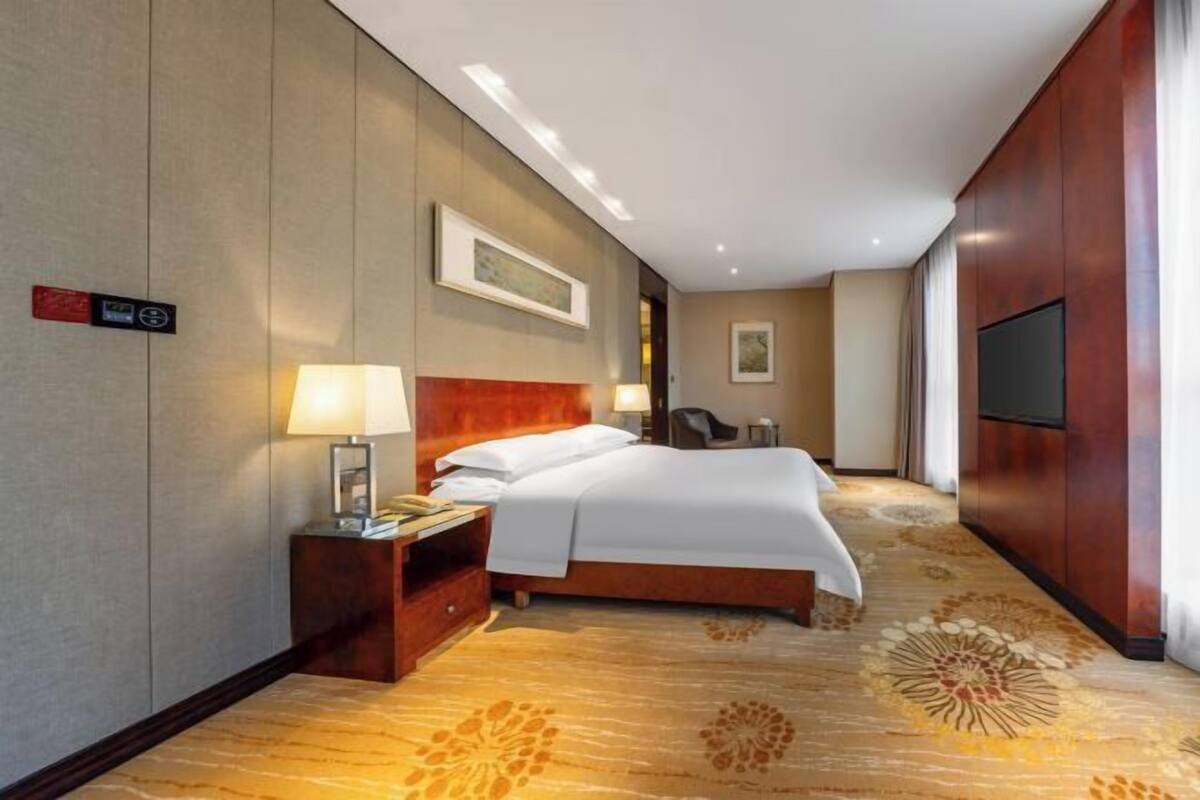 A hotel room with a bed, bedside table, lamp, artwork, and a television.