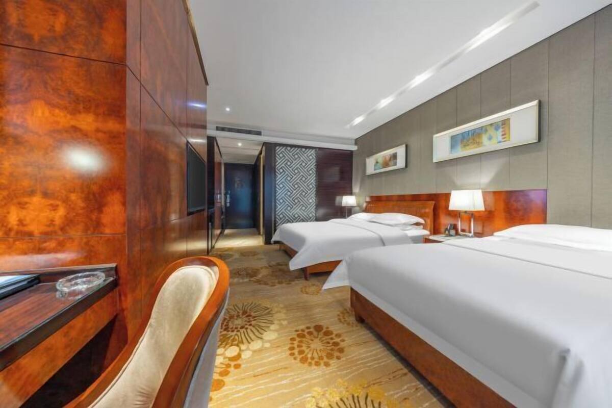 A hotel room with a wooden desk, two beds, a chair, and a patterned carpet.