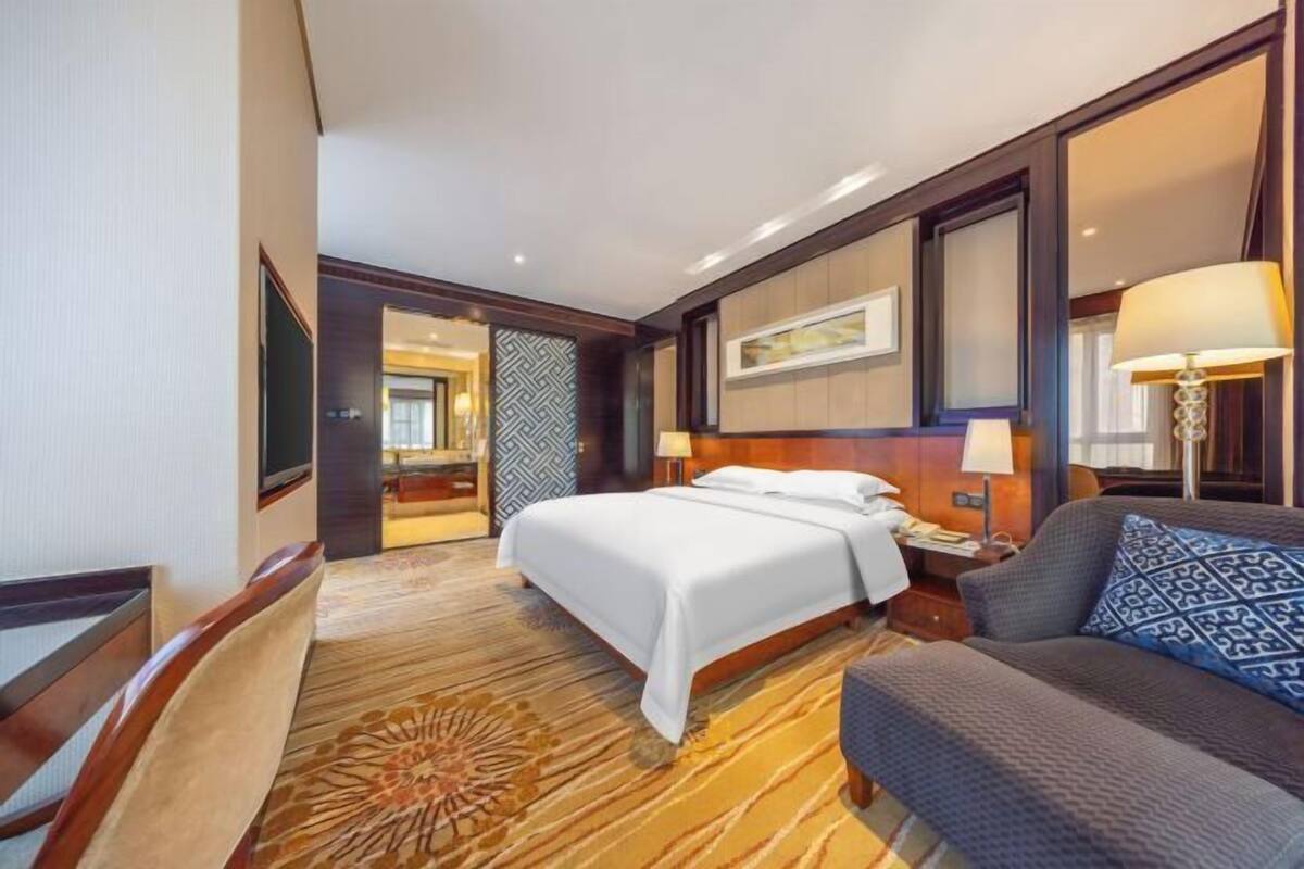 A hotel room with a bed, a sofa, a desk with a lamp, and a large mirror.