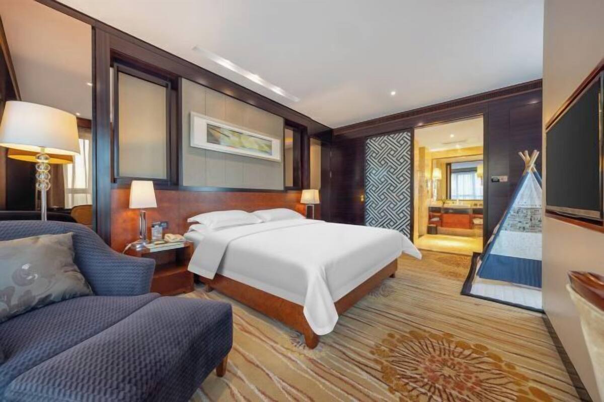 A hotel room with a bed, a sofa, a desk with a lamp, and a wall-mounted TV.