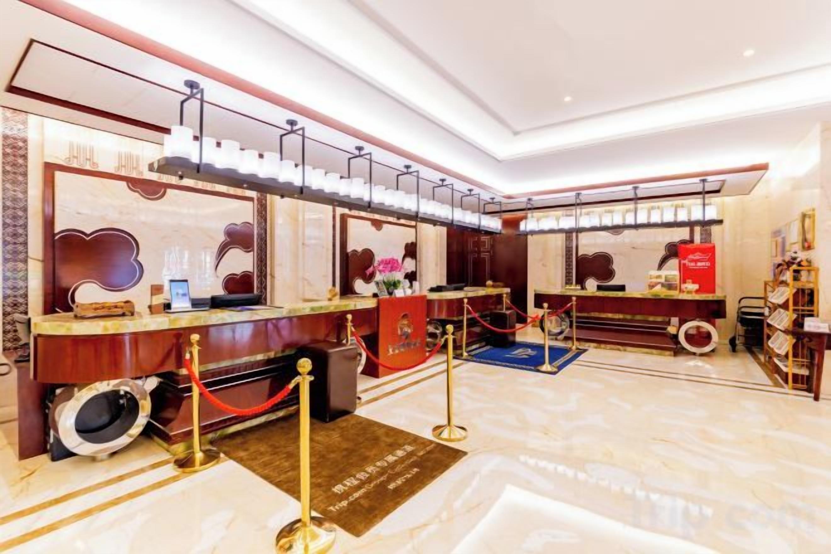 Área de recepção de hotel com piso de mármore, balcão de atendimento, tapete vermelho e corda de veludo delimitando a área.