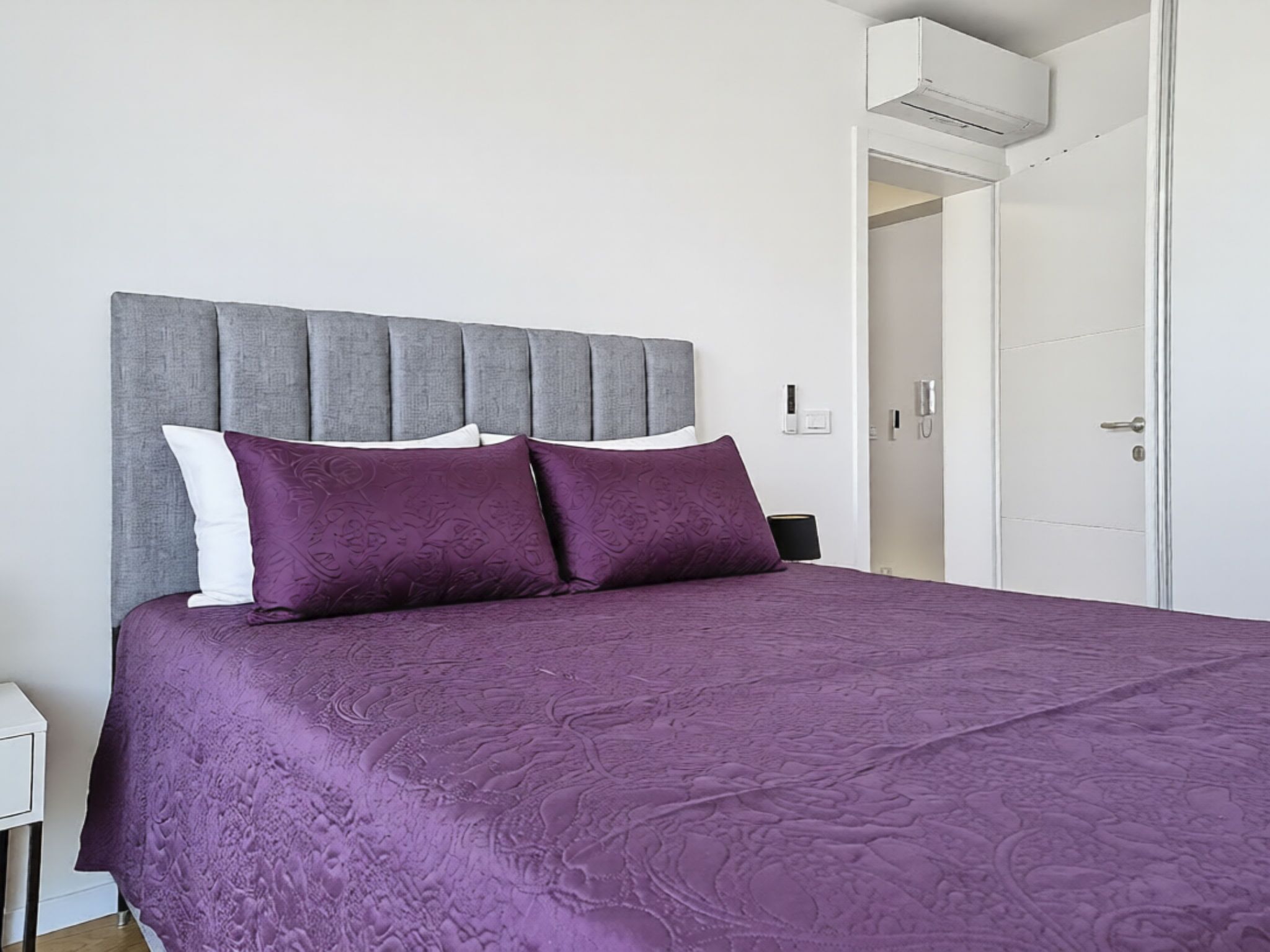 A neatly made bed with a grey headboard and purple bedding, a white nightstand, and a white door with a silver handle.