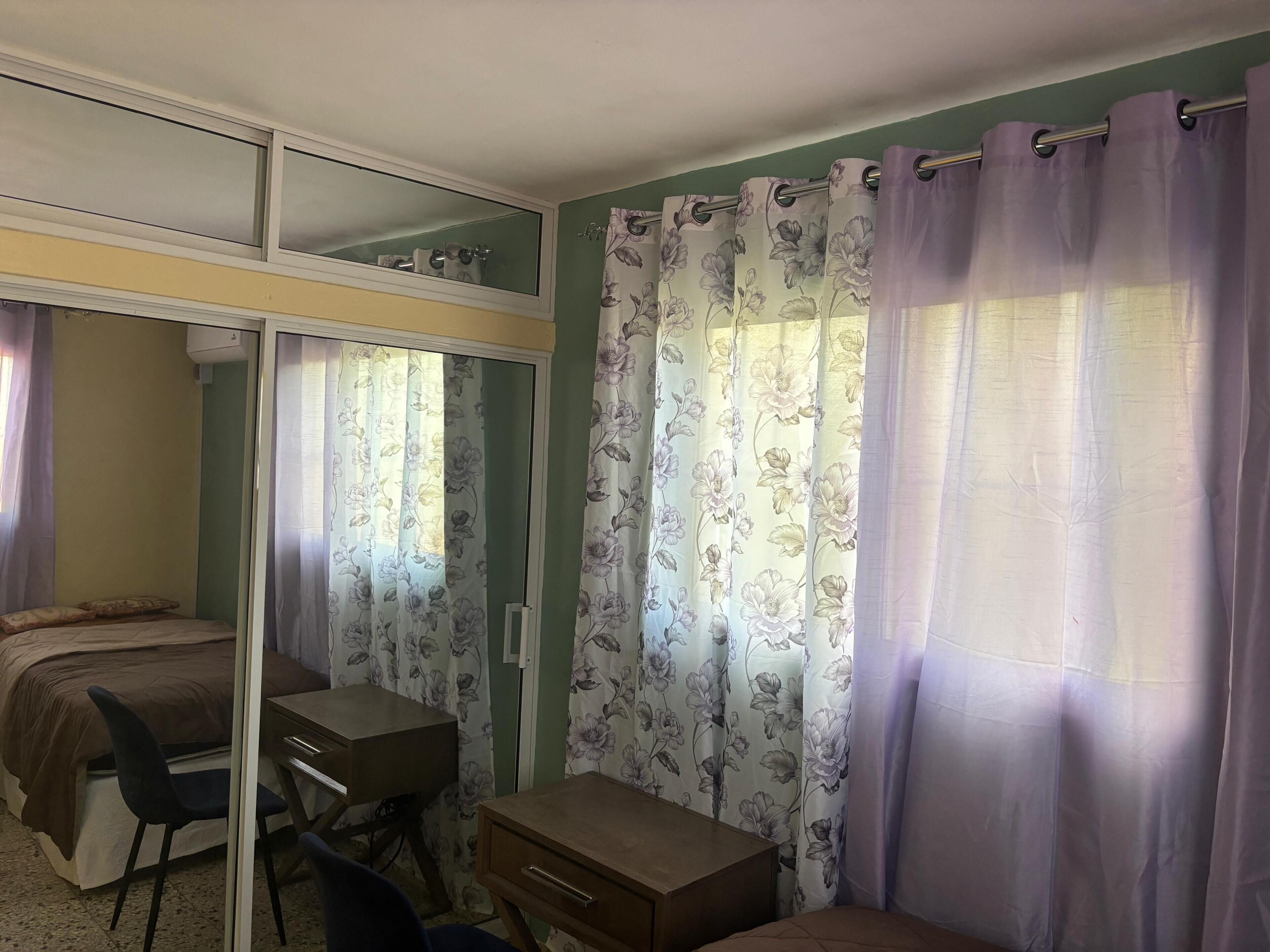 A bedroom with a bed, a desk, a chair, a mirror, and curtains.