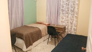 Une petite chambre avec un lit simple, un bureau, une chaise et deux rideaux.