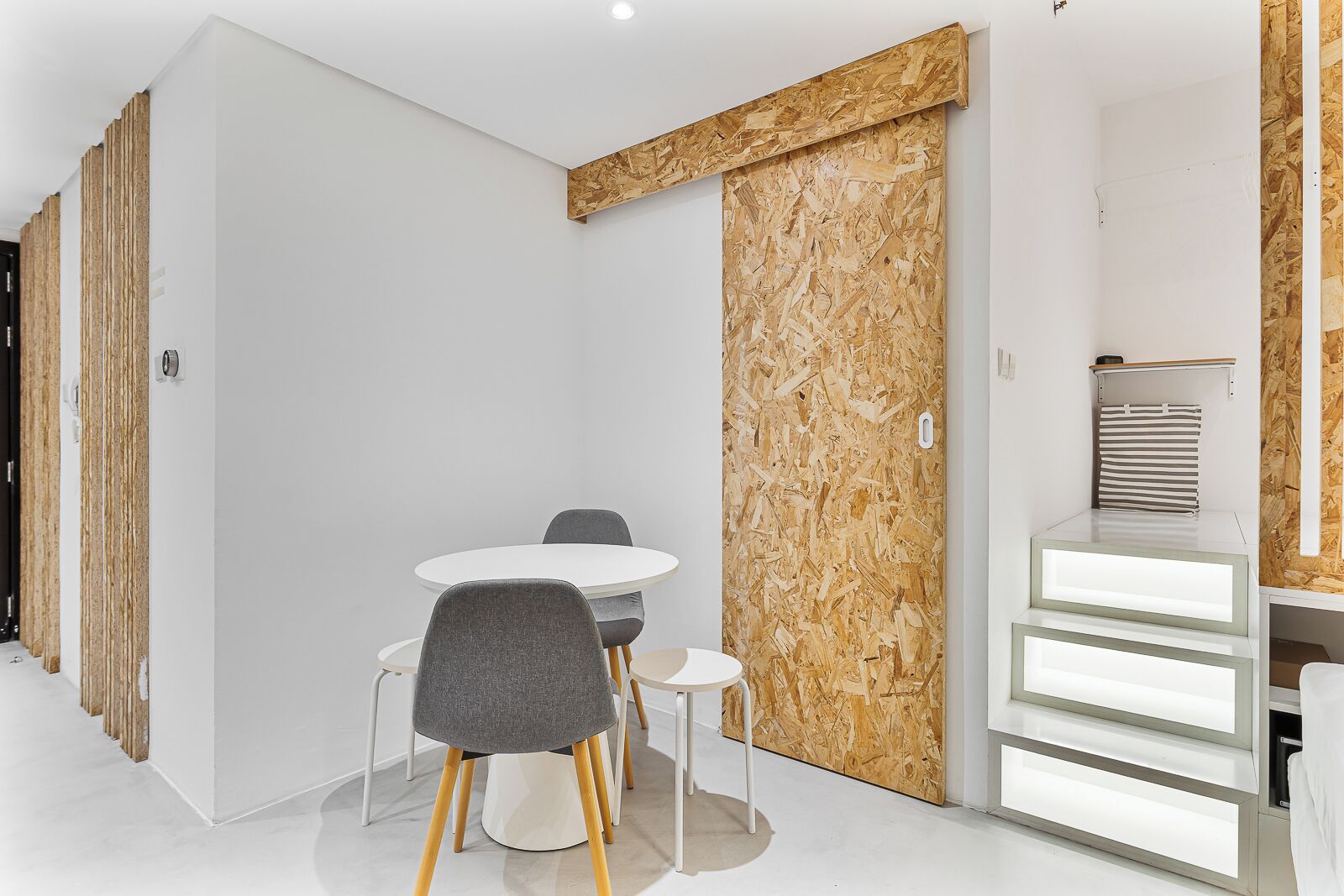 Ein modernes Zimmer mit weißer Wand, einer Wandverkleidung aus Holz, einem runden weißen Tisch mit zwei Stühlen und einer weißen Kommode mit Schubladen.