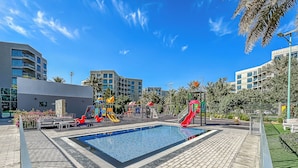 Una piscina con un tobogán rojo, rodeada de un solárium pavimentado y un parque infantil con diversas estructuras de juego.