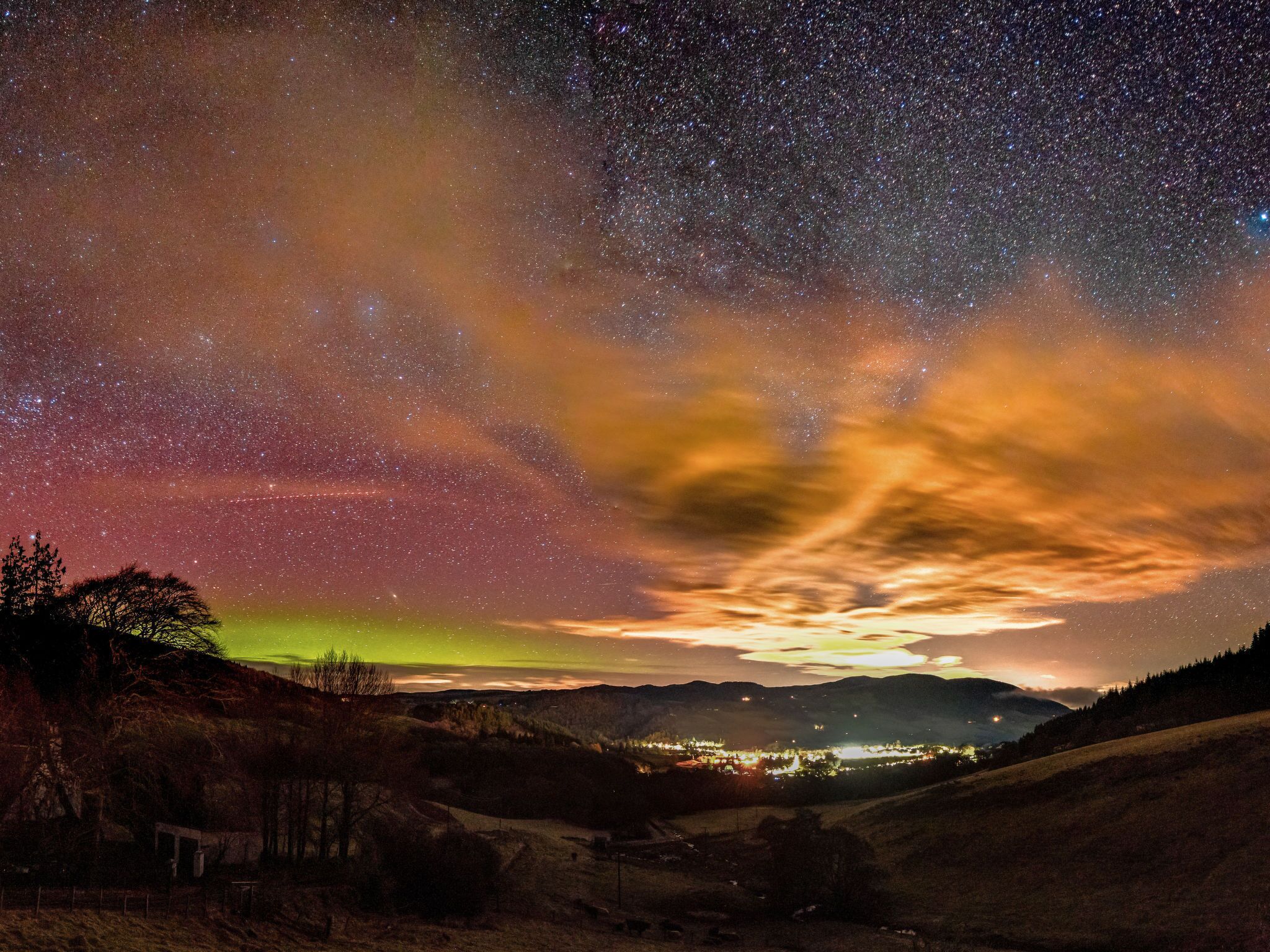 Cielo nocturno lleno de estrellas y una aurora boreal colorida sobre un valle con luces distantes.