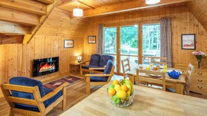 Ein gemütliches Blockhaus-Interieur mit Kamin, Holzmöbeln, einem Esstisch mit Obst und großen Fenstern.