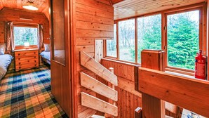 Eine Holzhütte mit Inneneinrichtung, darunter eine Treppe, ein Bett, eine Kommode und ein Fenster mit Blick auf Bäume.