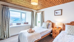 A bedroom with a wooden ceiling, a large window with curtains, a bed with a headboard, a bedside table with a lamp, and a framed picture on the wall. - Bunloit Combi by Interhome (Drumnadrochit)