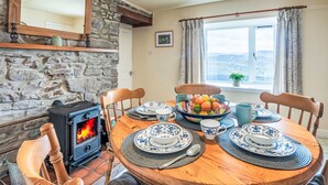 A cozy dining room with a stone wall, a wood-burning stove, and a table set for four with blue and white plates and a fruit bowl. - Bunloit Combi by Interhome (Drumnadrochit)