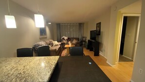 Un comedor con mesa y sillas de granito, un conjunto de sofás, un televisor y un pasillo que conduce a otra habitación.