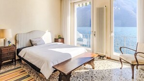 Ein Hotelzimmer mit einem Bett, einem Nachttisch aus Holz, einer Lampe, einem Sessel, einem Tisch und Meerblick vom Balkon.