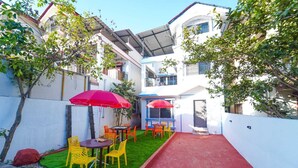A courtyard with a small lawn, outdoor seating with yellow chairs and red umbrellas, and a building with a white exterior and blue accents. - Ananya Holidays Villa (Lonavala)