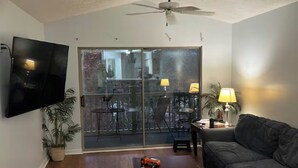 Una sala de estar con ventilador de techo, televisor de pantalla plana, sofá, mesa de centro y puerta corrediza de vidrio que da a un patio al aire libre.