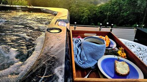 A hot tub with a wooden tray containing a plate with bread and a cup, set against a scenic view of a forested landscape. - Hot spa with Ocean Views - fire pit - 
2 fully equipped retreats - privacy  (Bonogin)