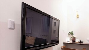 A flat-screen TV mounted on a wall, a wooden chair, a small table with a plant, and a light switch. - Asakusa The Apartment (Tokyo)