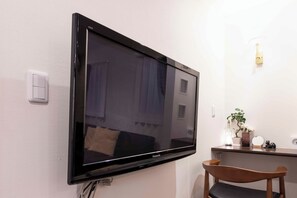 A flat-screen TV mounted on a wall, a wooden chair, a small table with a plant, and a light switch. - Asakusa The Apartment (Tokyo)