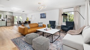 A modern living room with a sofa, ottomans, a coffee table, and a chair. There is a kitchen in the background with a refrigerator and cabinets. - Cozy Cabin! BBQ • Spa • Fire Pit • Kids Play Zone (Mariposa)