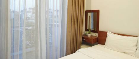 Une chambre d’hôtel avec un lit, une table de chevet avec un miroir, une plante en pot et des rideaux légers.