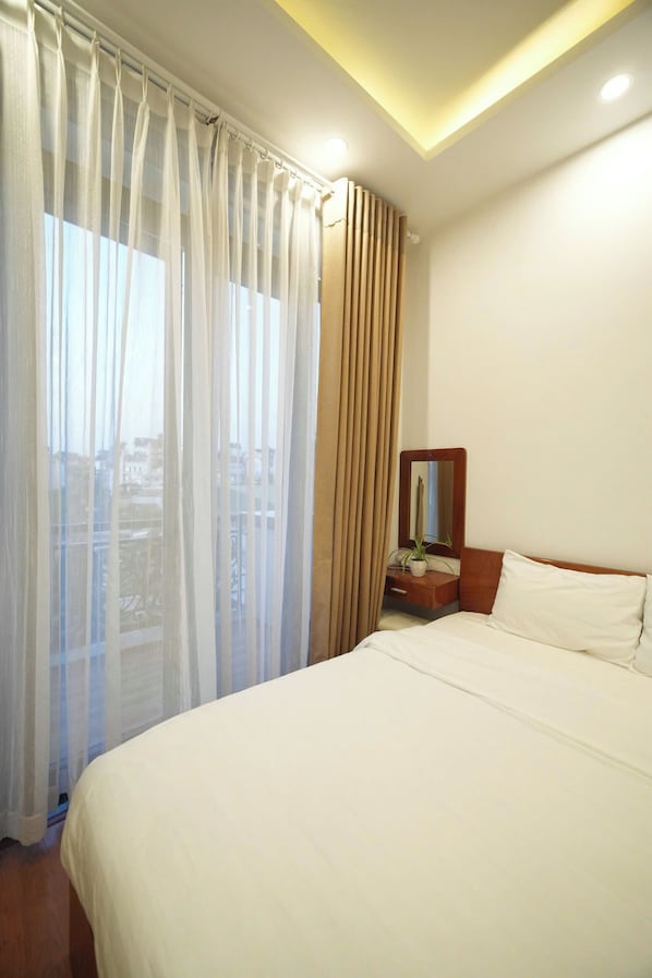 Une chambre d’hôtel avec un lit, une table de chevet avec un miroir, une plante en pot et des rideaux légers.