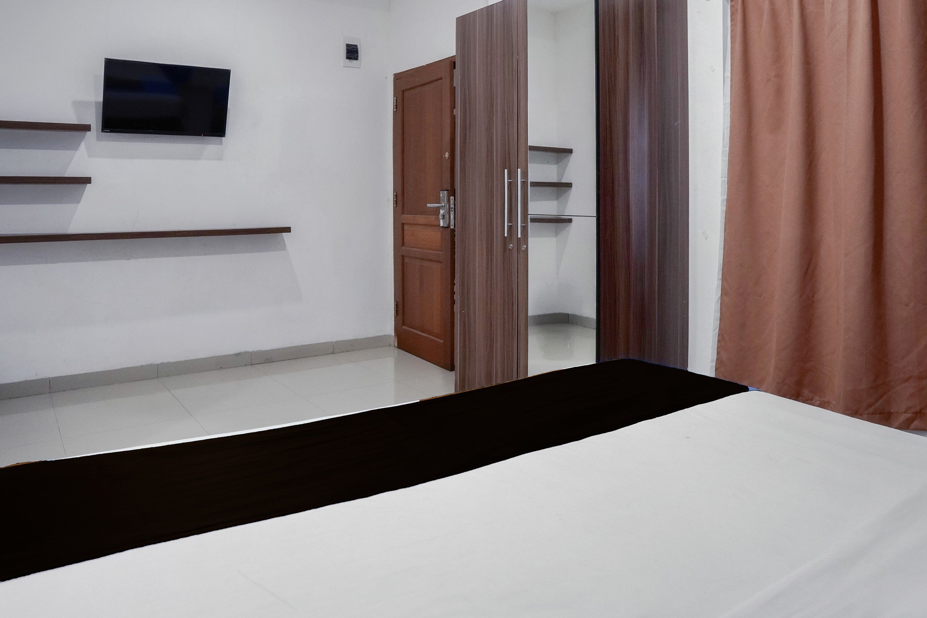 A bedroom with a bed, a TV mounted on the wall, a wardrobe, and a door leading to another room.