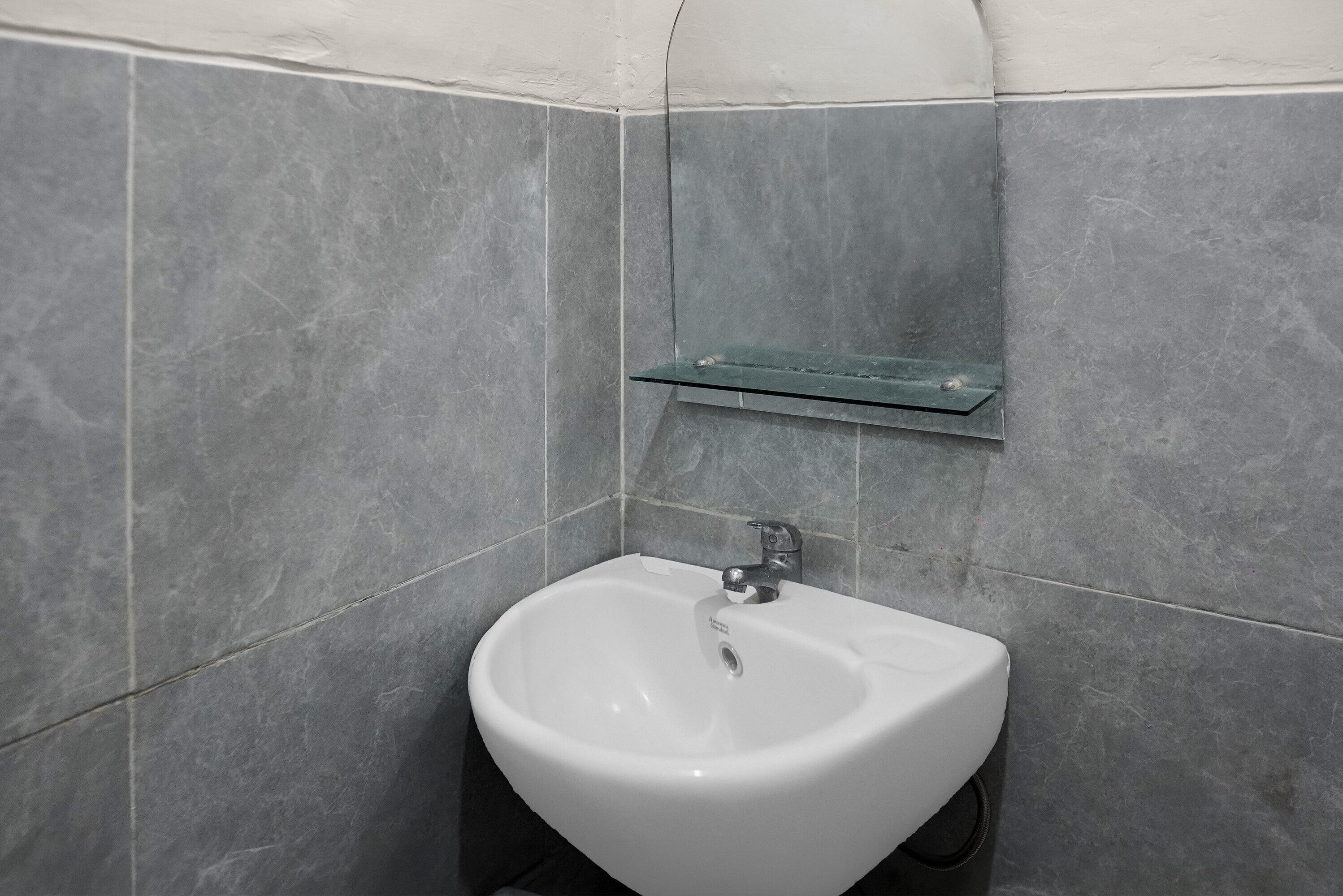 Een badkamer met een witte wastafel, grijze tegels en een klein plankje erboven.