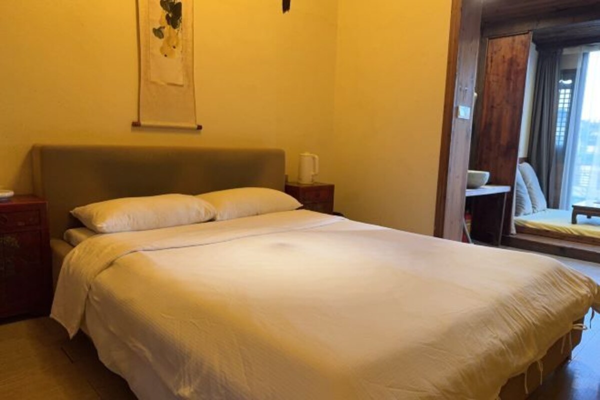 A hotel room with a large bed, a wooden headboard, a bedside table with a cup, and a window with a view.