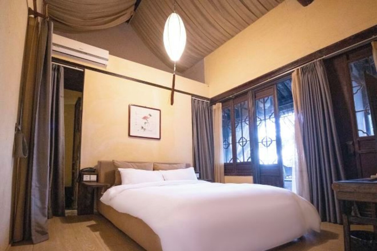 A bedroom with a bed, a bedside table, a nightstand, a window with curtains, and a ceiling light fixture.