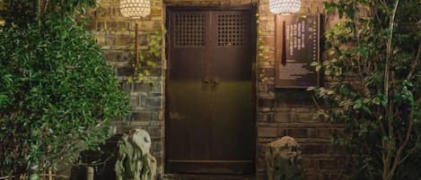 A stone entrance with a dark door, surrounded by greenery and two wall-mounted lights.