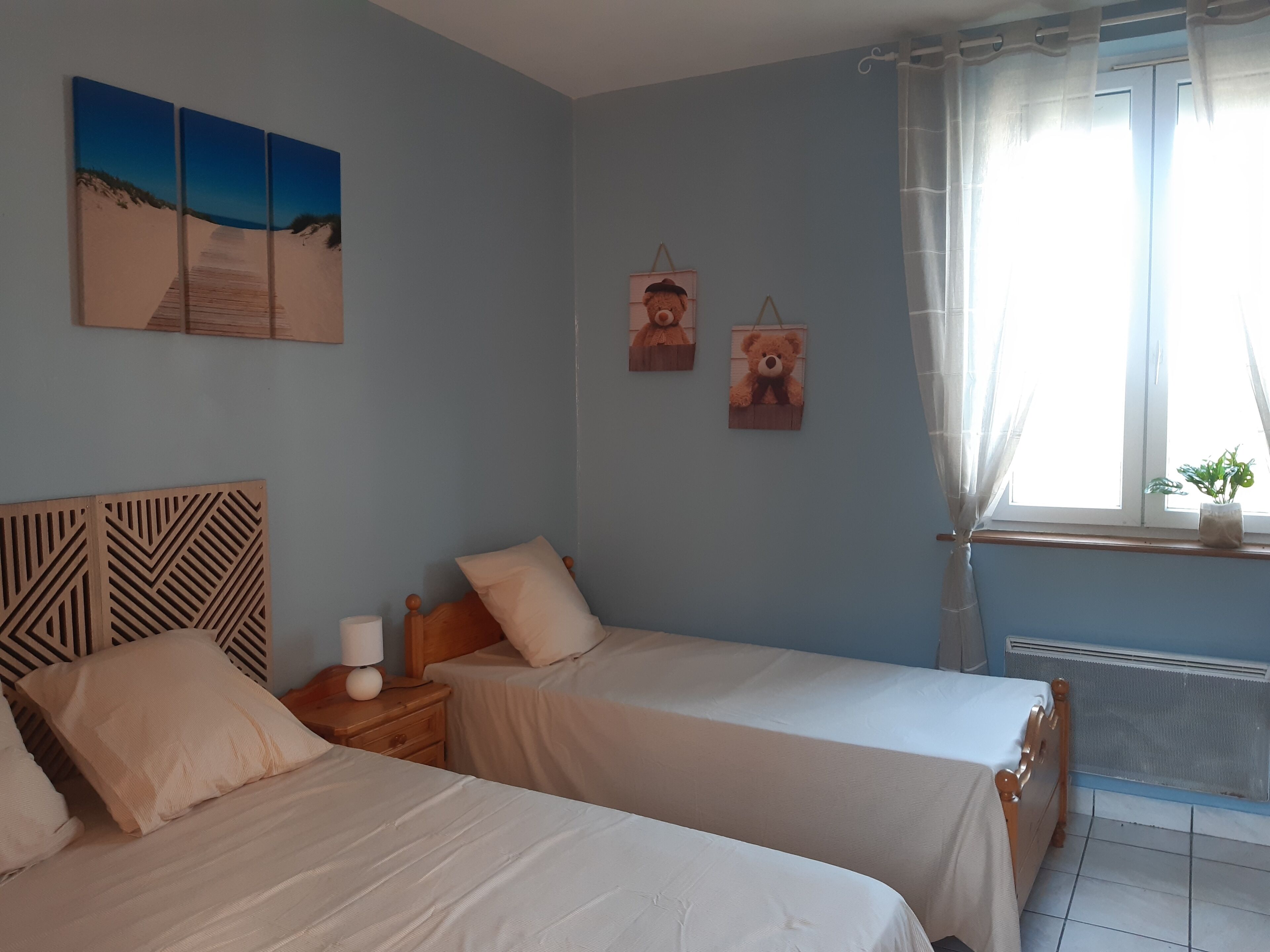 Une chambre avec deux lits, une tête de lit en bois, une table de chevet, une lampe, une fenêtre avec des rideaux et des œuvres d’art murales.