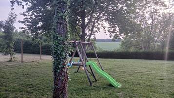 Un parque infantil con columpios y un tobogán, rodeado de césped y árboles.