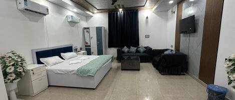 A modern hotel room with a bed, a sofa, a TV, and an air conditioning unit.