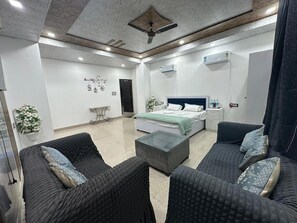 A modern hotel room with a bed, two sofas, a coffee table, a ceiling fan, and an air conditioning unit. - The Luxe Haven (Sonepat)