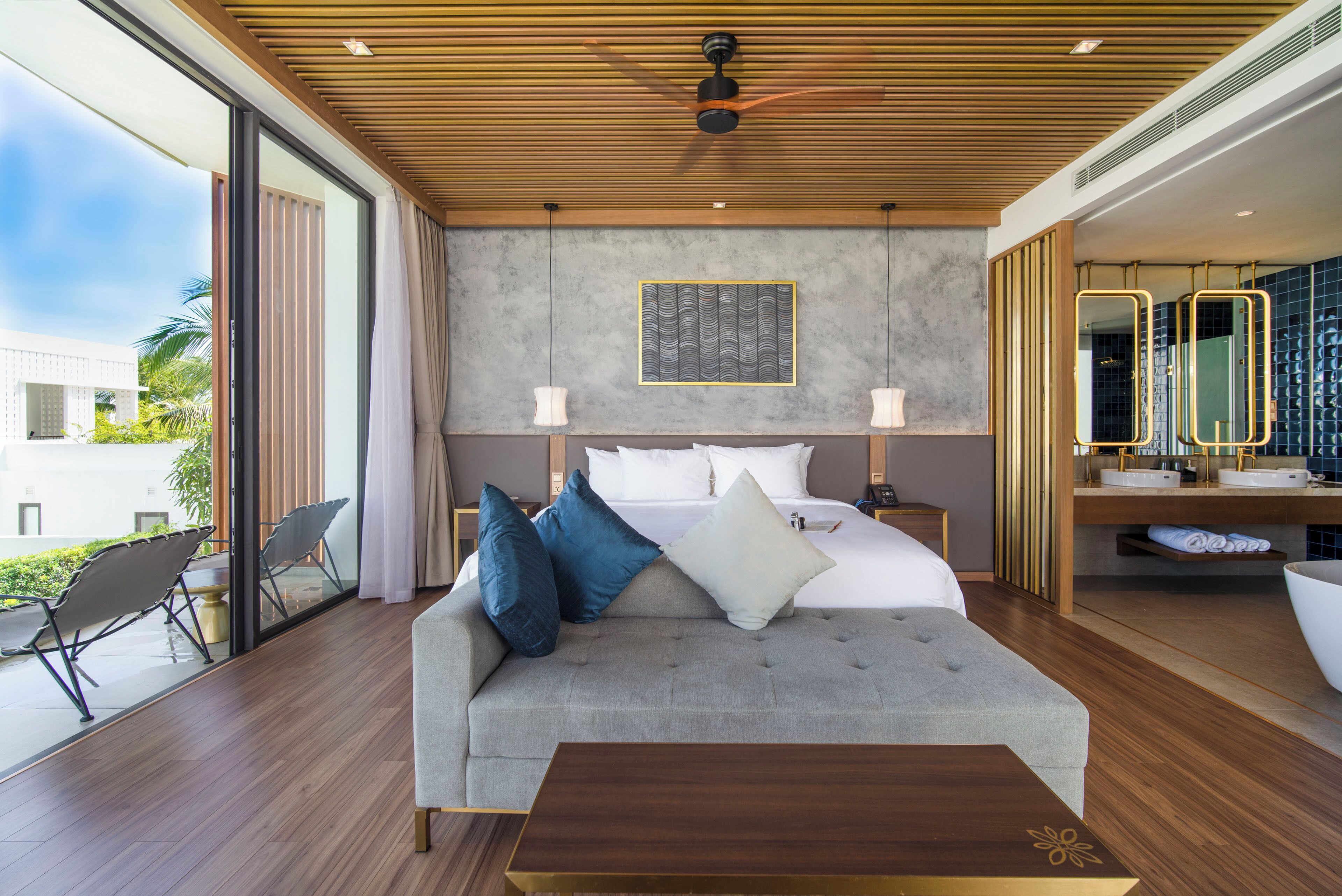 A modern hotel room with a large bed, a sofa, a wooden ceiling, and a view of the outdoors.