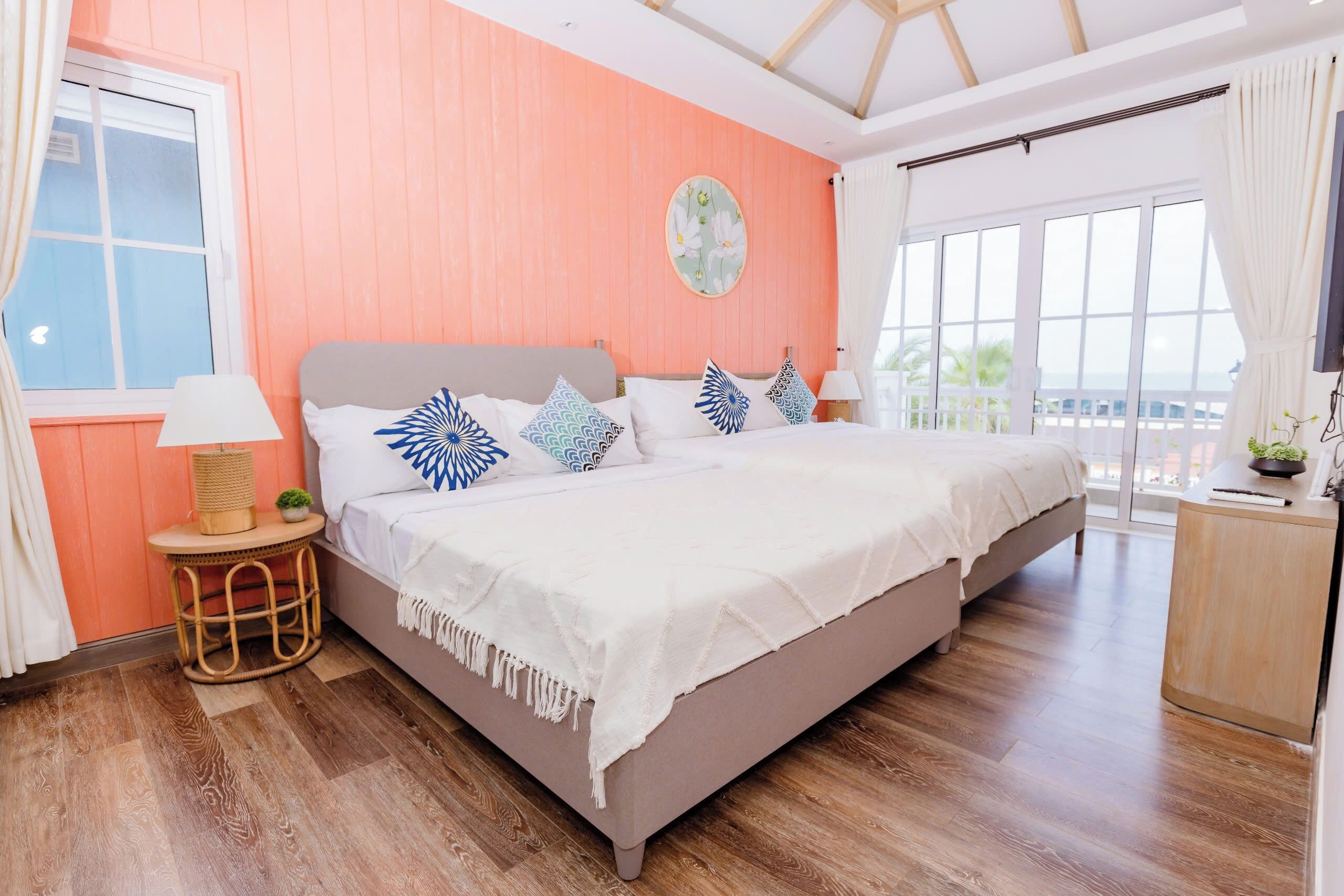A bedroom with a bed, a round table with a lamp, a wall clock, and a window with curtains.