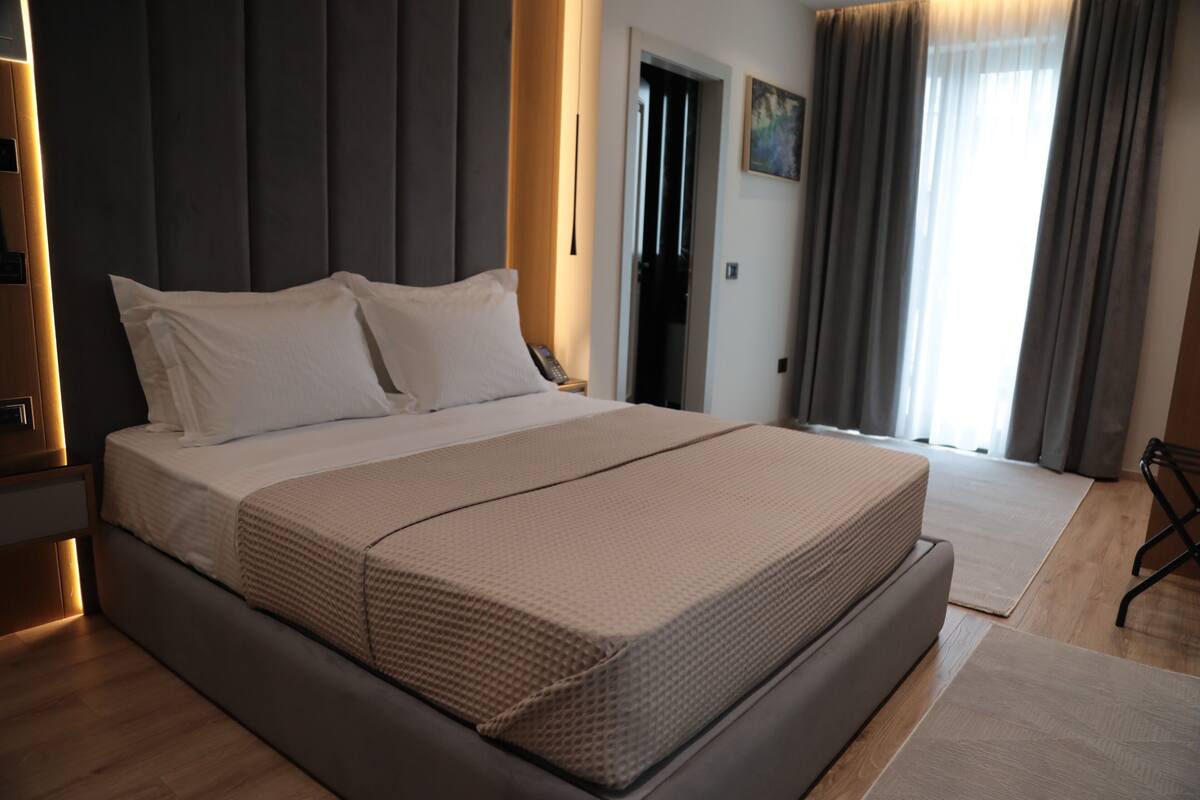 A modern hotel room with a large bed, a bedside table, a nightstand, a door, and a window with curtains.