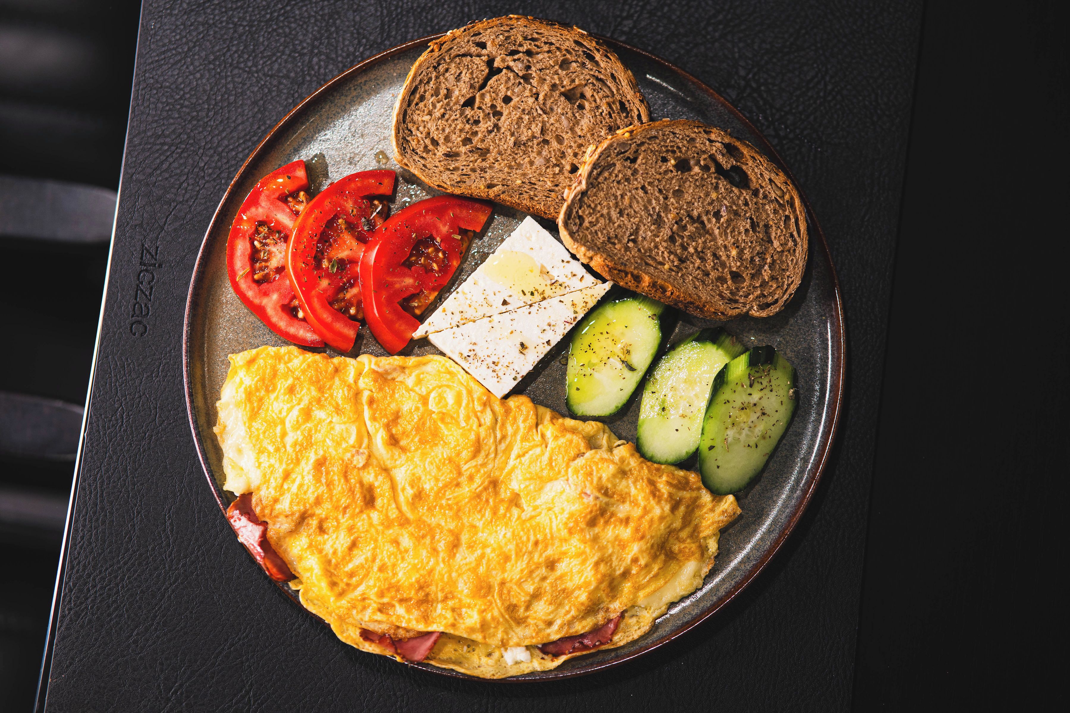 Une assiette de petit-déjeuner comprenant une omelette, des tranches de tomate, du fromage, des rondelles de concombre et deux tranches de pain complet.