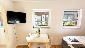 A room with a wooden floor, a dining table with chairs, a bed with a quilt, a TV mounted on the wall, and two windows with potted plants. - Apartment "Prague" (Korschenbroich)