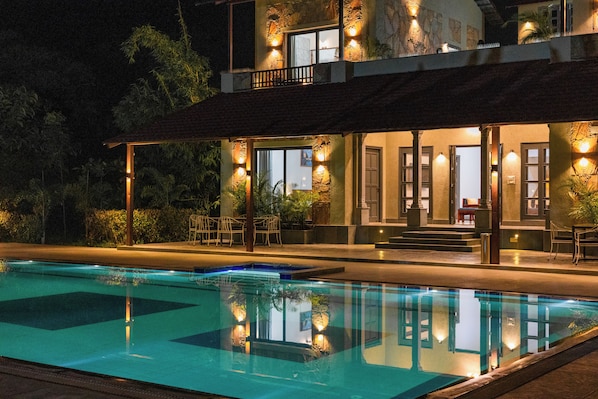 A poolside view of a hotel with a pool, outdoor seating, and a balcony with lights.