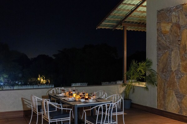 Un espace extérieur aménagé pour les repas, avec une table dressée pour quatre personnes, entourée de chaises blanches, un mur en pierre et une vue sur le ciel nocturne.