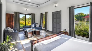 Habitación de hotel moderna con cama, sofá y zona de comedor, ventanas amplias con cortinas y vistas a zonas verdes.