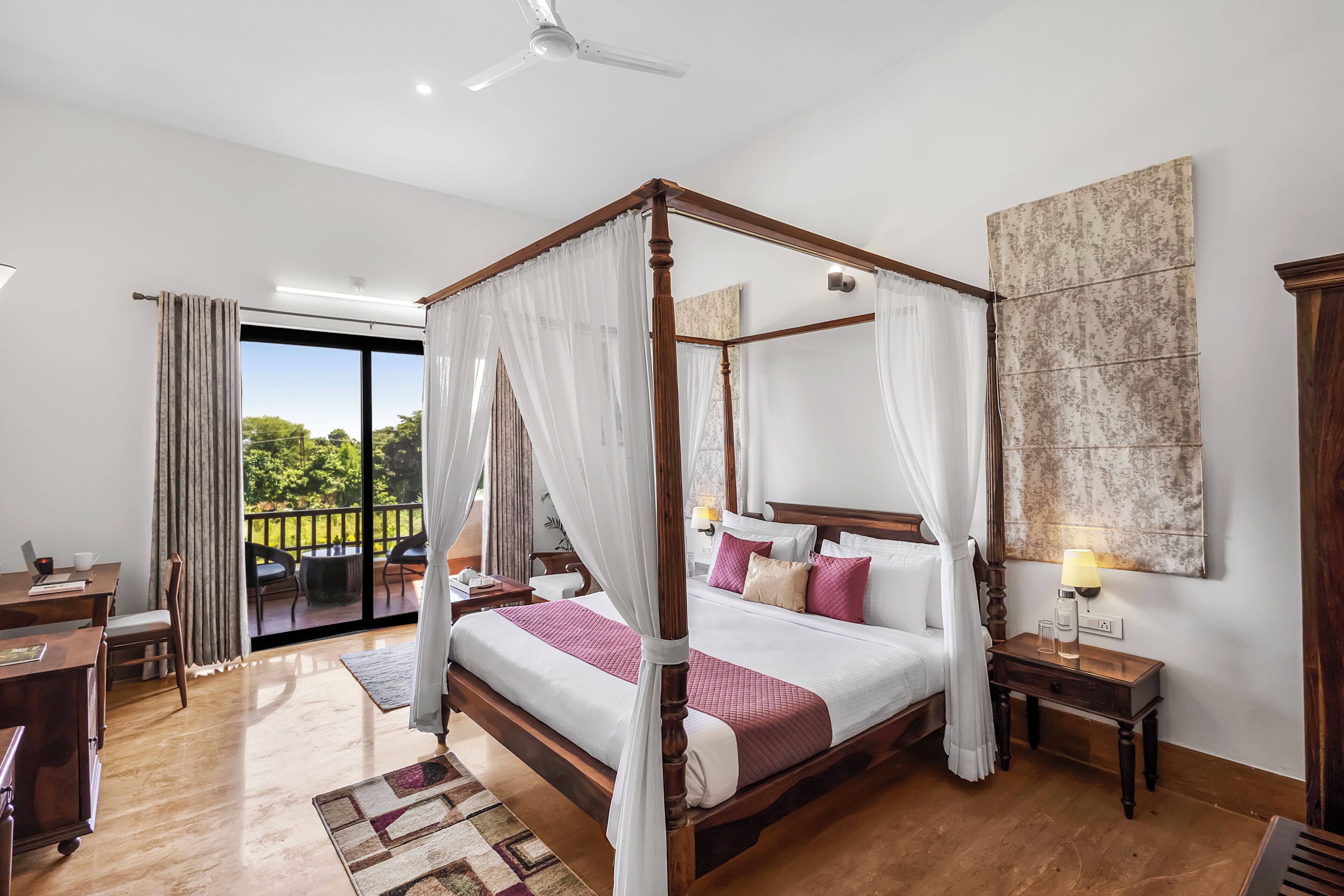 A four-poster bed with a canopy, a wooden nightstand, a desk with a chair, and a view of the outdoors.