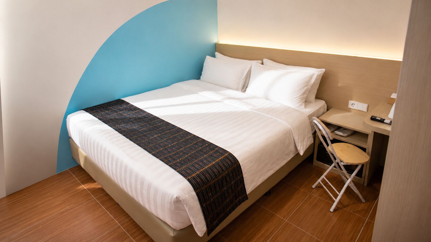 A neatly made bed with a white and black striped blanket, a small desk with a chair, and a wall-mounted light.