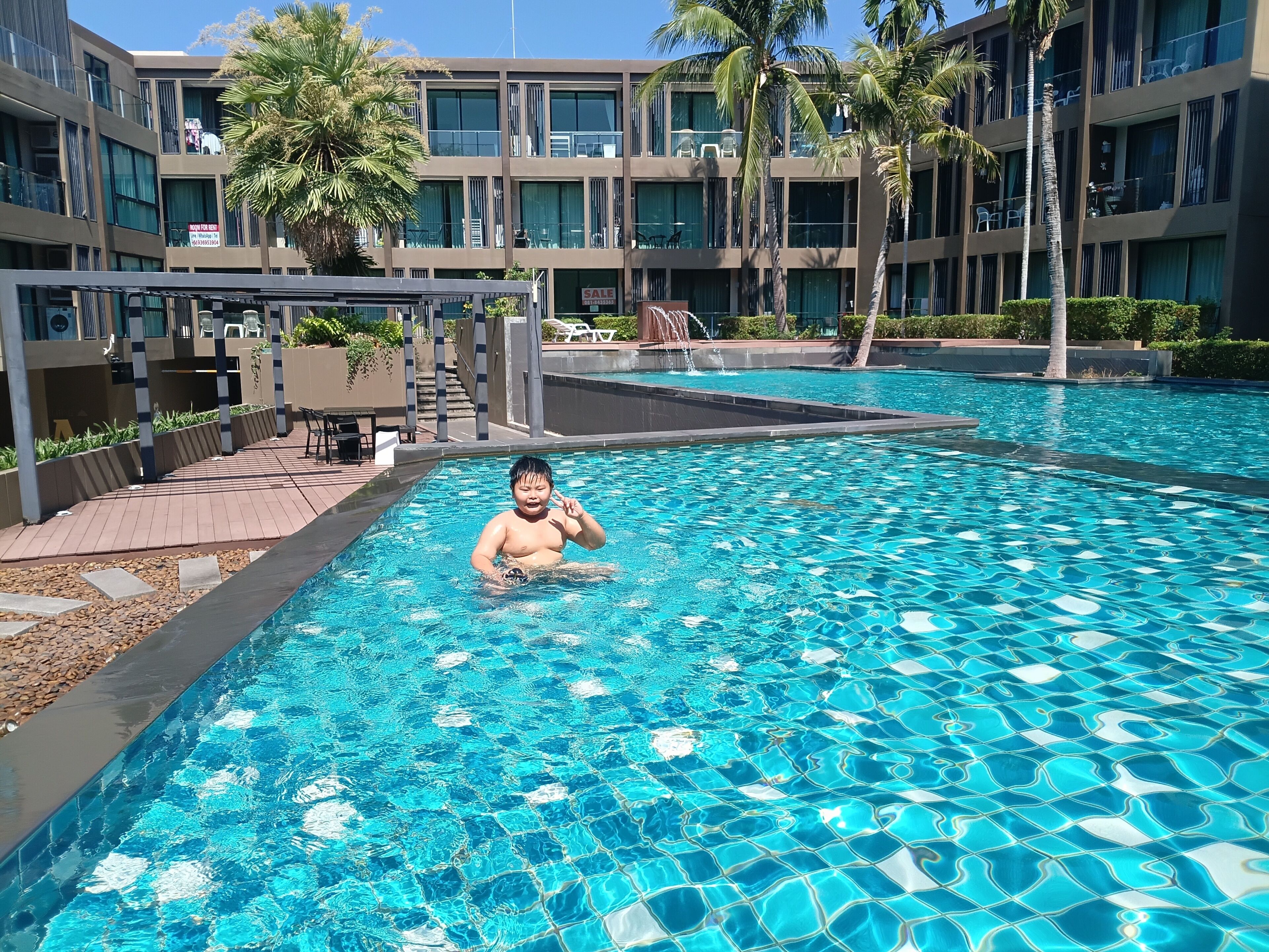 A swimming pool with clear water, surrounded by a modern building with balconies and palm trees.