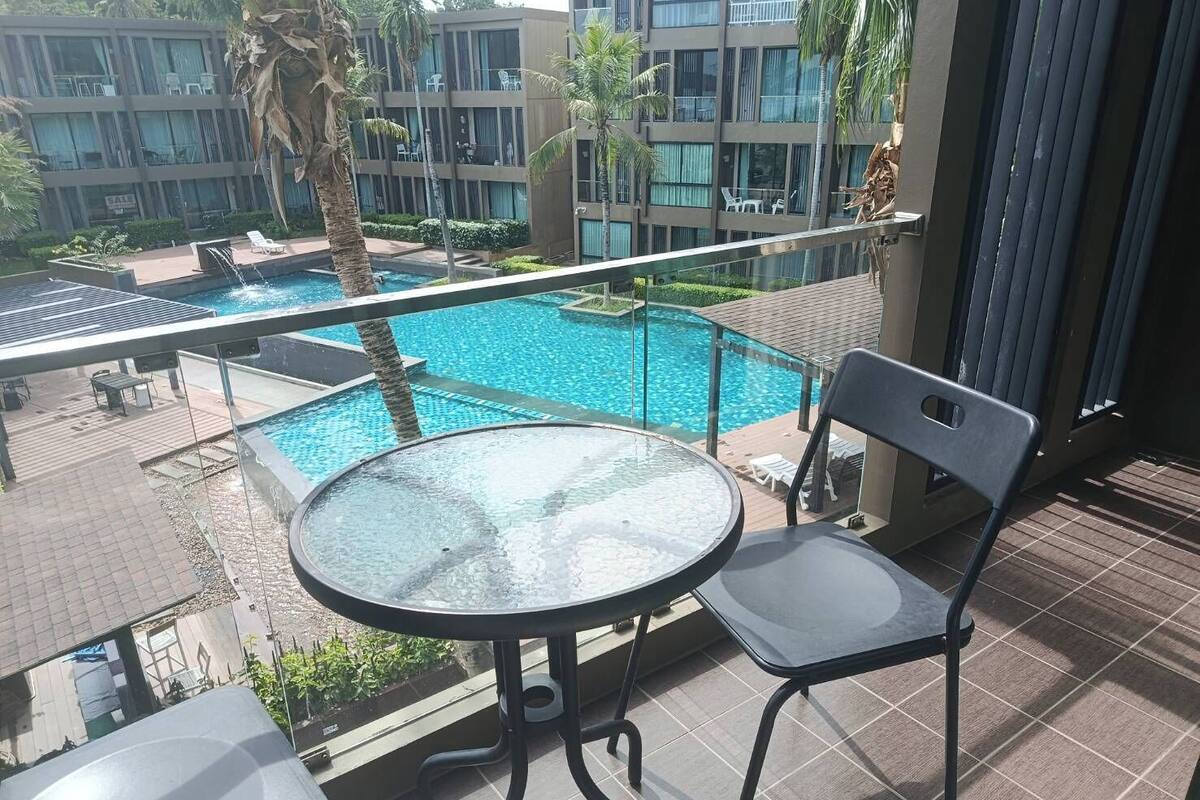 Un balcon doté d’une table et d’une chaise en verre, offrant une vue sur une piscine et sur un immeuble à plusieurs étages, lui-même agrémenté de balcons.