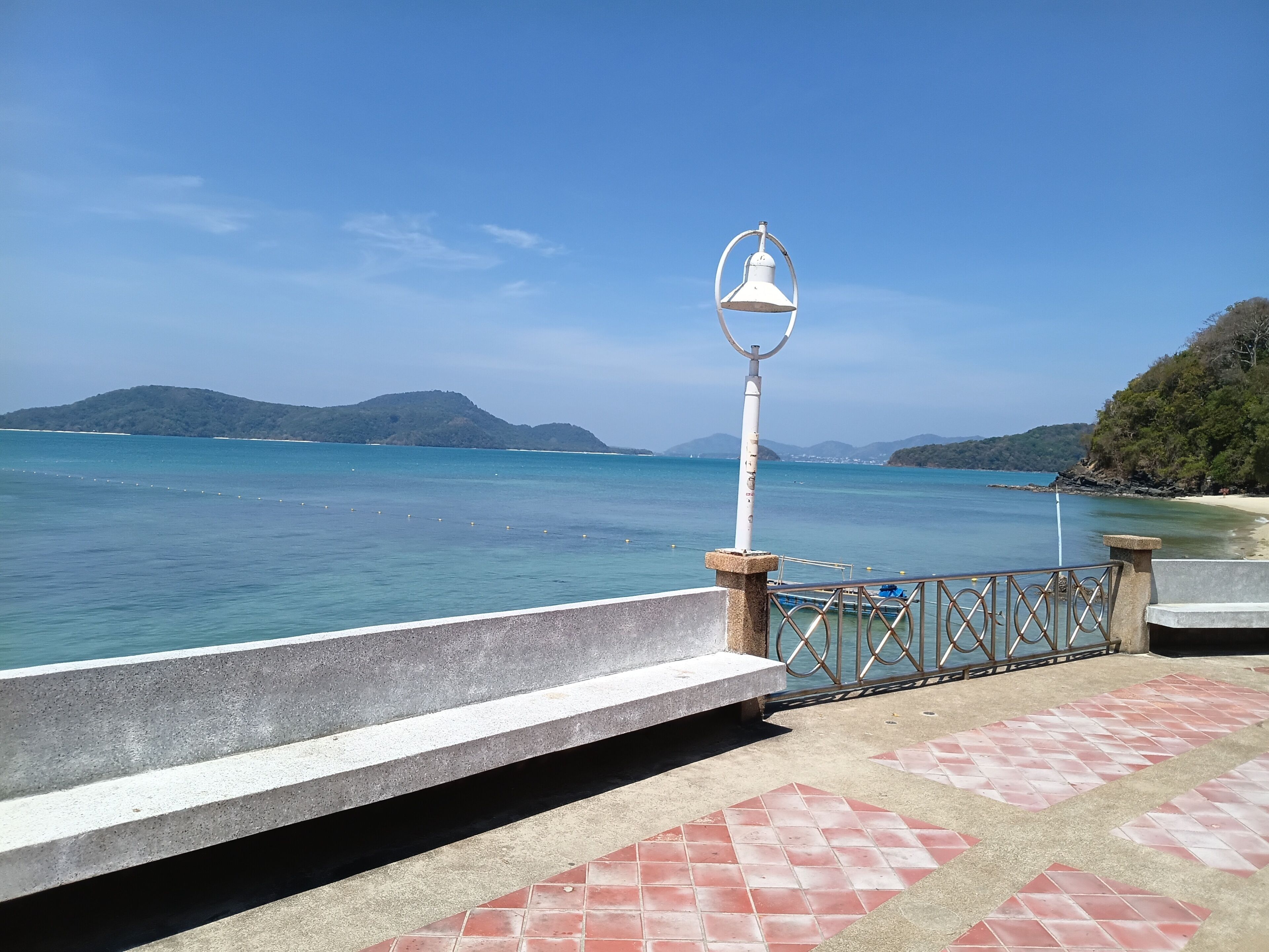A coastal walkway with a metal railing, a white lamp post, and a bench.
