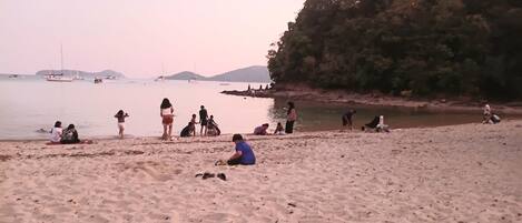 A sandy beach with people sitting and standing, boats in the water, and a forested area in the background.
